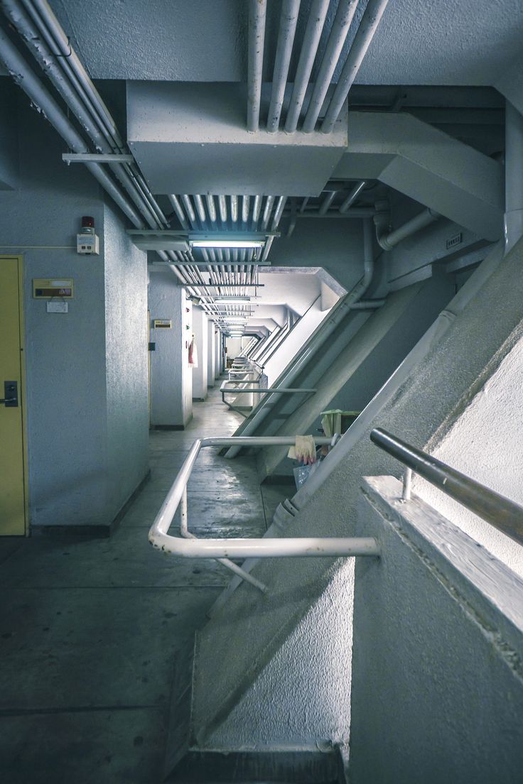 the image shows a long, narrow corridor or hallway in a building. the architecture is industrial in style, with exposed metal pipes and concrete structures. the walls are a muted grey color.  there are stairs visible further down the hallway, and the perspective creates a sense of depth. the overall feel is cold and somewhat austere. a section of railing is prominent in the foreground.