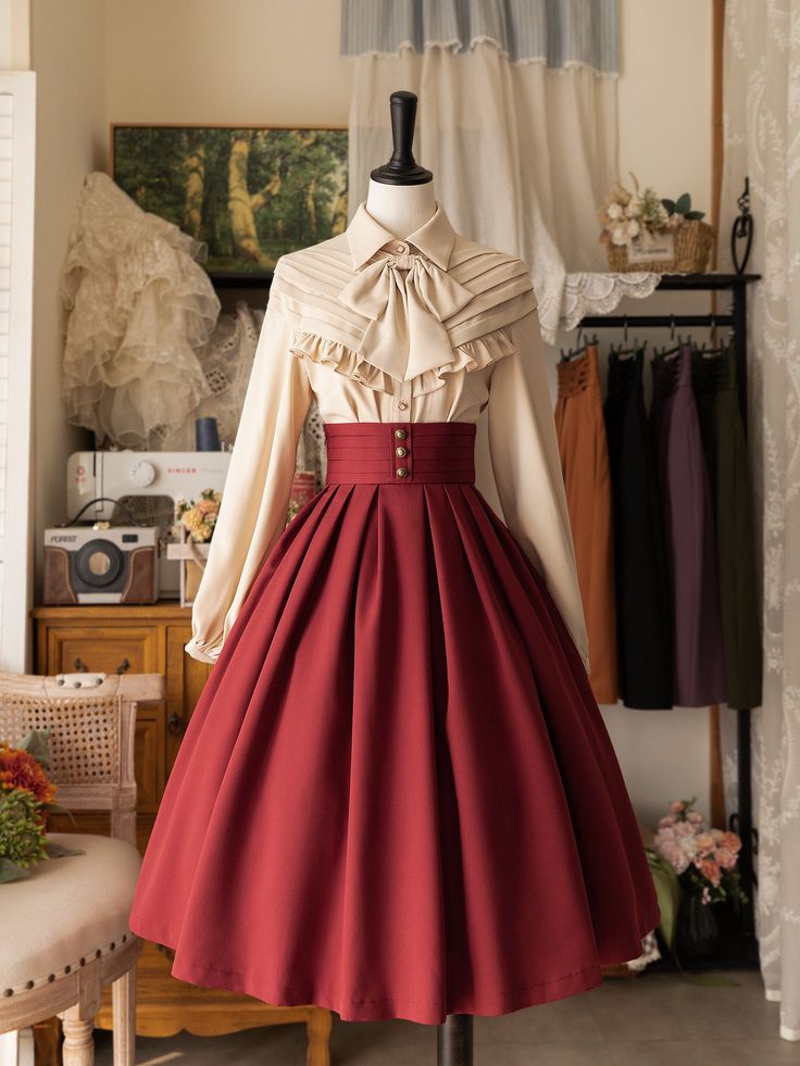 a mannequin displays a long vintage style dress composed of a highwaisted burgundy skirt and a longsleeve cream blouse. the blouse has a large collar with a bow tied at the neck and is tucked into the skirt. the skirt falls to the floor in full pleats and has a row of buttons down the front. the top button is fastened. the mannequin is posed in front of a white curtain and a wooden cabinet. to the right of the mannequin is a black clothing rack with brown and black fabric hanging from it. the overall style of the dress and setting is romantic and vintage.
