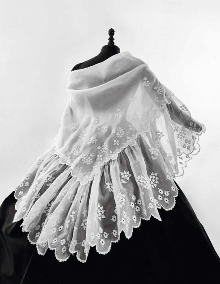 the image shows a closeup of a delicate white lace shawl draped over a black dress on a mannequin.  the shawl is sheer and features intricate embroidery with a floral motif and a scalloped edge. the lace appears to be handmade or antique, suggesting a vintage or possibly even historical piece of clothing. the black dress provides a strong contrast to the white lace, making its detail stand out. the overall feel of the photograph is elegant and refined, likely intended to highlight the beauty and craftsmanship of the lace shawl.  the monochrome presentation further emphasizes the texture and details of the garment.