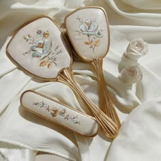 two vintage, embroidered, hand mirrors with gold handles laying on a white background. the background has an elegant drape to it. the mirrors are ornate and feature beautiful embroidered floral designs. the overall image is elegant and feminine.