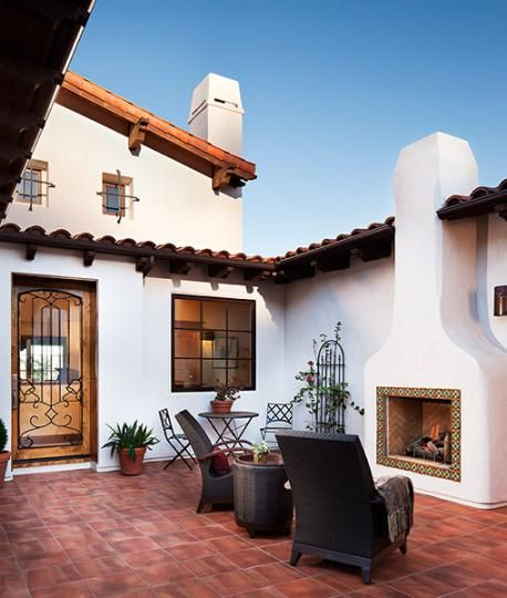 an outdoor patio is surrounded by white stucco walls with clay tile roofing. two chairs are positioned near a fireplace that has a decorative tile border. potted plants add greenery, and a wrought iron door is visible to the left. a small table with chairs sits near a window.