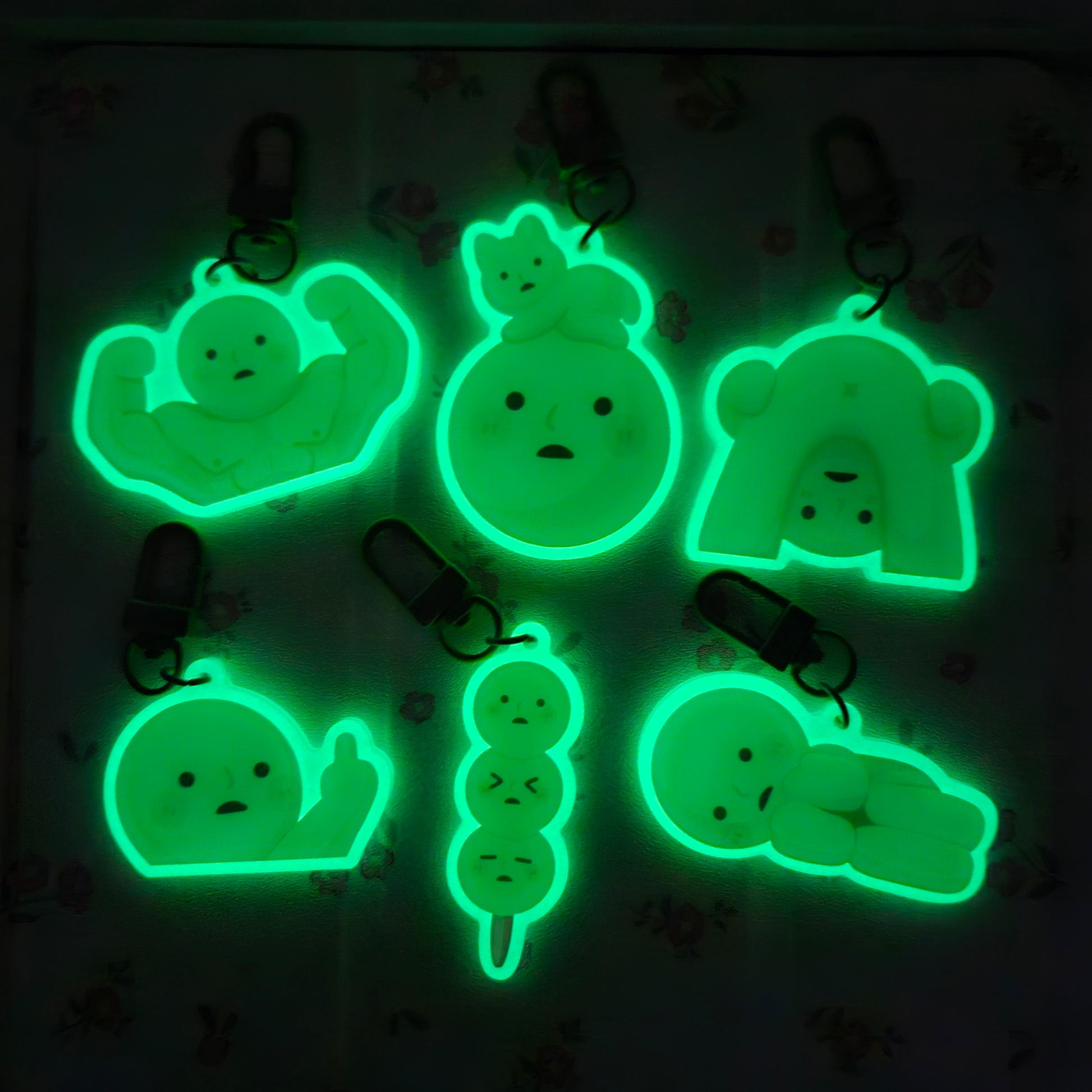 the image shows a collection of eight glowinthedark keychains. each keychain features a different small cartoon character with various expressions, including a muscle man, a sadfaced ball, a relaxed ball with a cat sitting on it, a grumpyfaced dango (japanese sweet treat), a small person, and a bearlike character.  the keychains are illuminated in a dark environment, showcasing their glowinthedark properties. they appear to be made of a translucent material like plastic or acrylic.  the overall style is cute and chibilike.