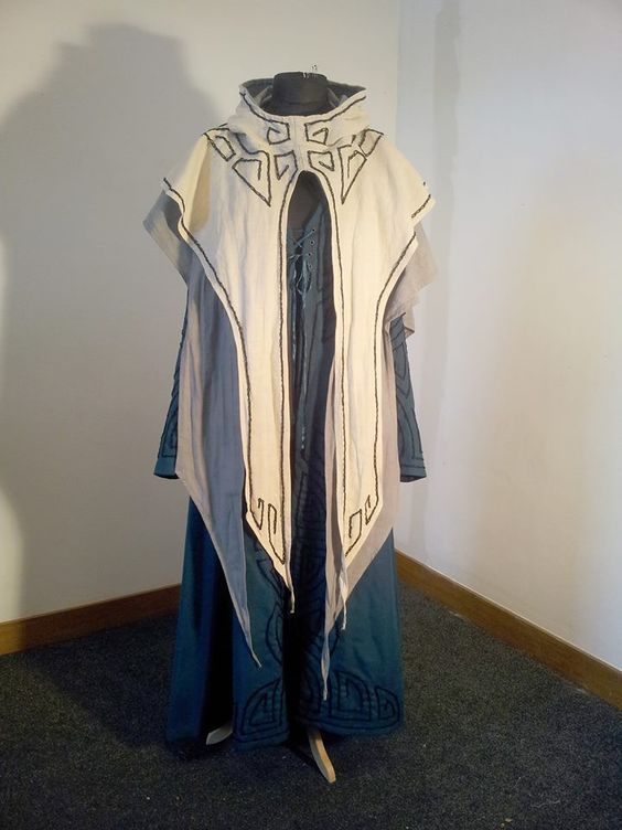 a long blue robe with a white hooded overlay, the  overlay has an intricate black geometric pattern sewn around the edges. the bottom of the robe has black geometric patterns that run down the skirt.  the robe is on a dress form.