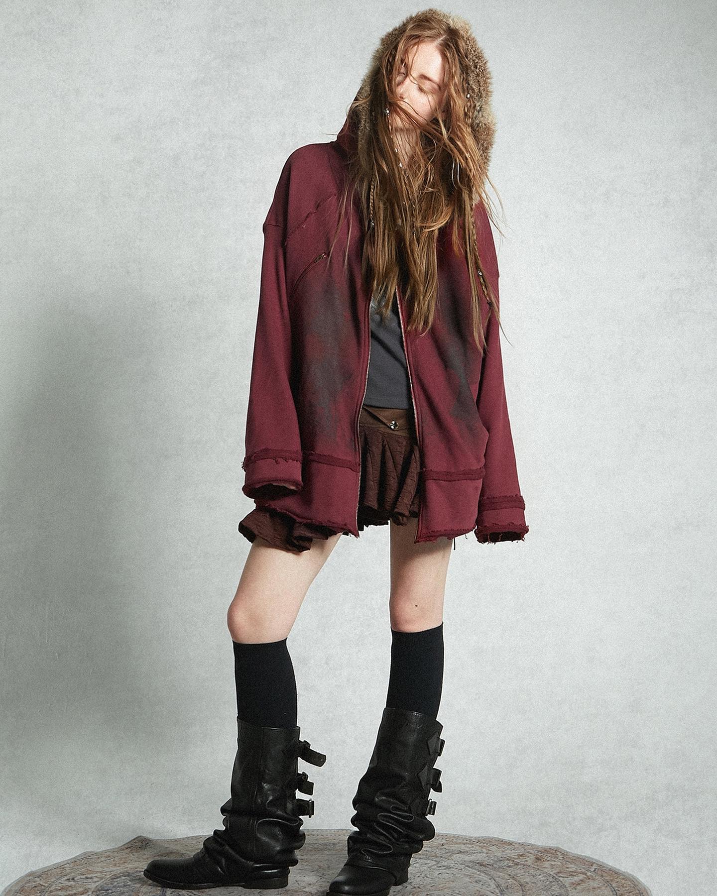 a fullbody shot of a female model showcasing a maroon oversized distressed hoodie with a furtrimmed hood, paired with a brown pleated mini skirt, black kneehigh socks, and black high boots with buckles. the model is posing in a studio setting against a light grey background. her long brown hair is slightly tousled. the overall style is grunge and vintageinspired, suitable for autumn or winter.