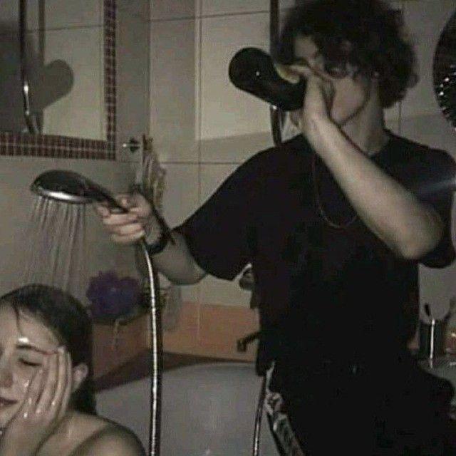 a woman is sitting in a bathtub with water running from a showerhead over her head. a person stands next to the tub drinking from a dark bottle. the scene is lit by dim lighting in a bathroom.
