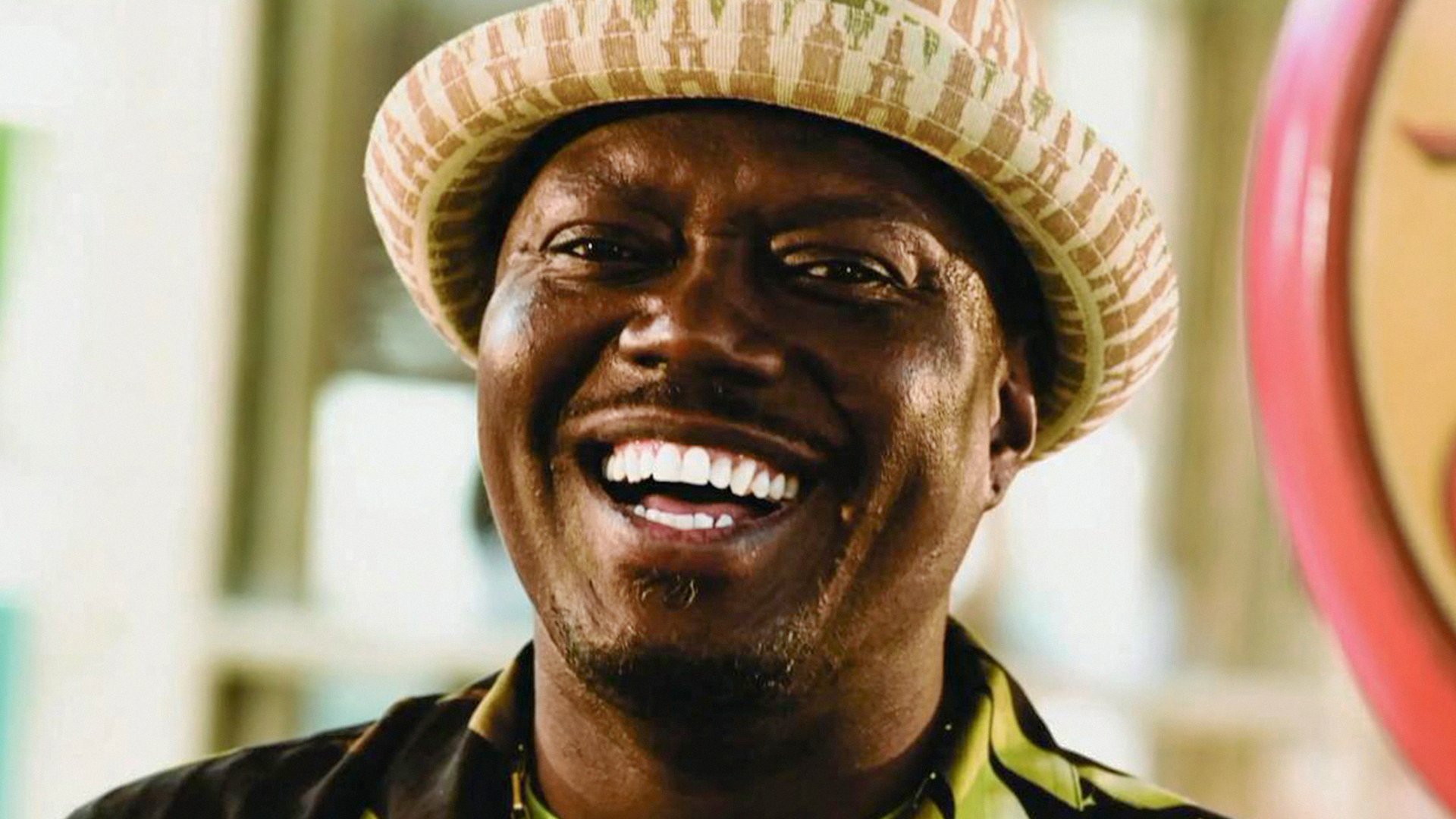 a closeup portrait of a black man wearing a straw hat and smiling broadly. he is looking directly at the camera and his teeth are visible. his expression is one of joy and happiness. the background is blurred and out of focus.