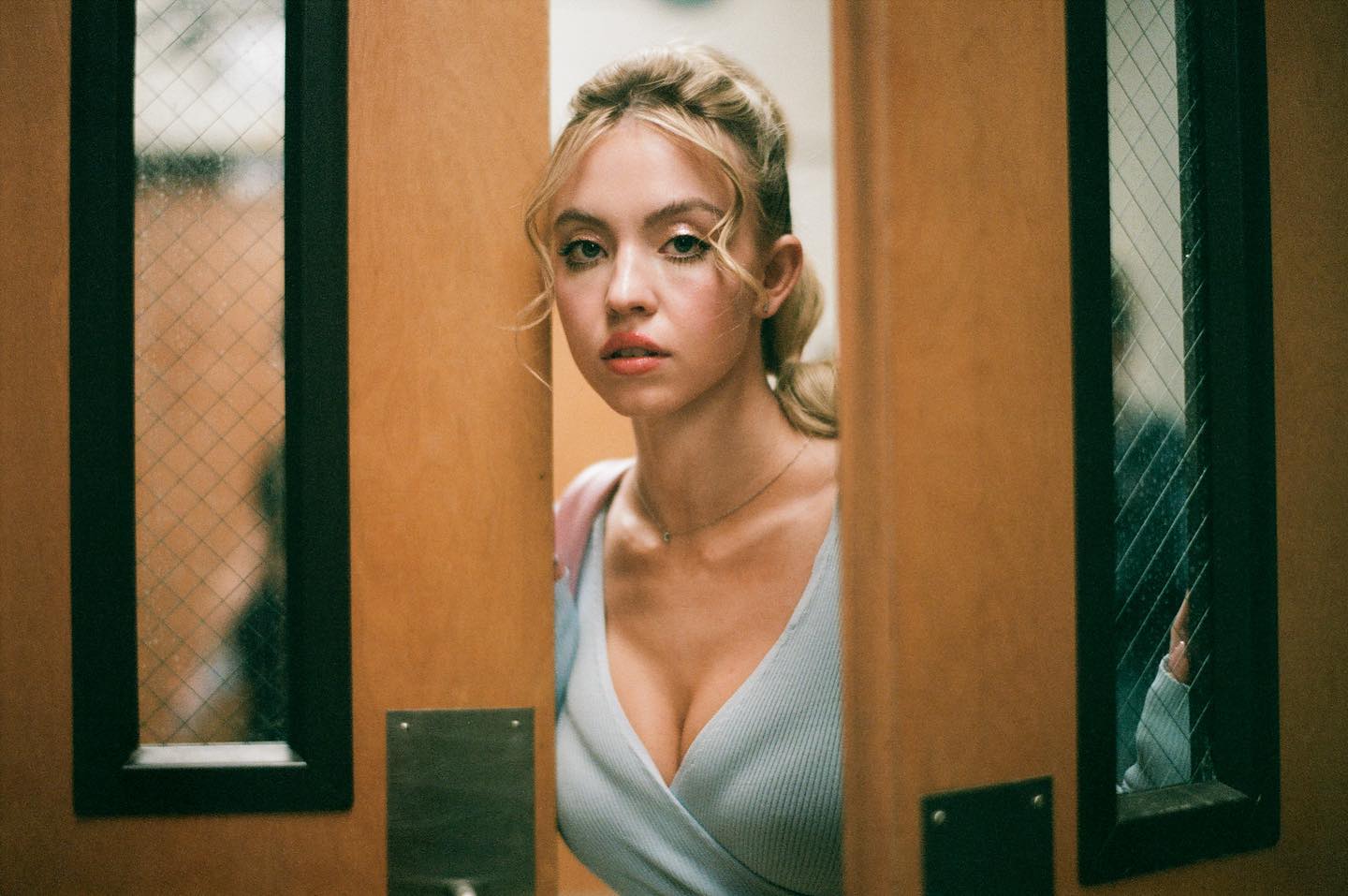 a young, attractive blonde woman with light makeup peeks through a doorway. she is wearing a light blue, ribbed vneck top that reveals some cleavage. the setting appears to be a school hallway or classroom, with the wooden door and a mesh window visible. the image has a shallow depth of field, focusing on the woman's face.  the overall feel is filmlike, natural, and a bit mysterious.