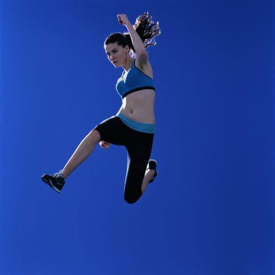 a woman with long brown hair pulled back is photographed jumping in the air against a bright blue sky. she is wearing black and turquoise running clothes and running shoes. her expression is determined, focused straight ahead. the sun highlights her and accentuates her muscles as she leaps.