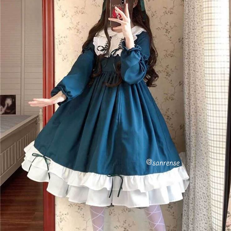 a young woman is standing in front of a mirror, taking a selfie. she is wearing a teal and white lolita fashion dress with long sleeves, a peter pan collar, and a lacetrimmed skirt. the dress has a bow at the neckline and ribbon detailing. the woman is wearing white tights and her hair is styled in two braids. the background is a bedroom with a bed and a mirror. the lighting is bright and artificial.