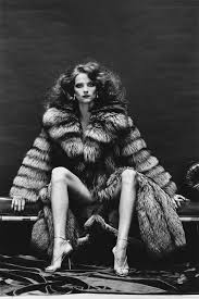 a black and white, full shot of a woman, possibly a model, posing in a luxurious, long fox fur coat. she is seated with her legs crossed, and her pose is both elegant and sensual. she's wearing highheeled strappy sandals. the background is dark and simple, focusing all attention on the woman and her attire. the overall style suggests a 1970s vintage aesthetic.