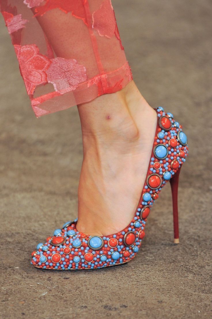 a close up shot of a woman's leg wearing red heels. the shoes are covered in red and blue gemstones. the woman is wearing a pink, floral dress. the picture is taken in a studio setting, with a gray background.
