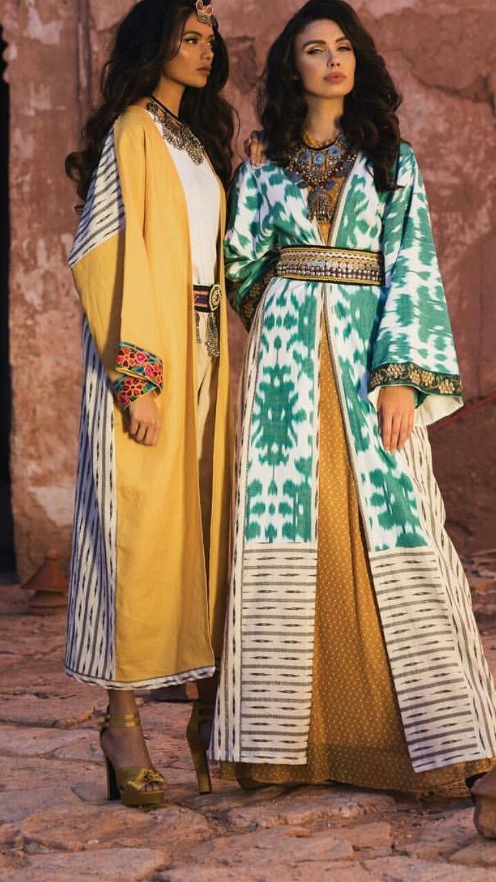 wo women in long, elegant dresses stand outdoors against an ancient stone wall. the woman on the left wears a mustardyellow abayastyle dress with offwhite and dark blue stripes at the sleeves. the woman on the right wears a teal and offwhite ikat pattern abaya with a golden belt. both are adorned with gold and gemstone jewelry, and are wearing gold highheeled sandals.  the setting suggests a historical or cultural photoshoot showcasing traditional clothing.