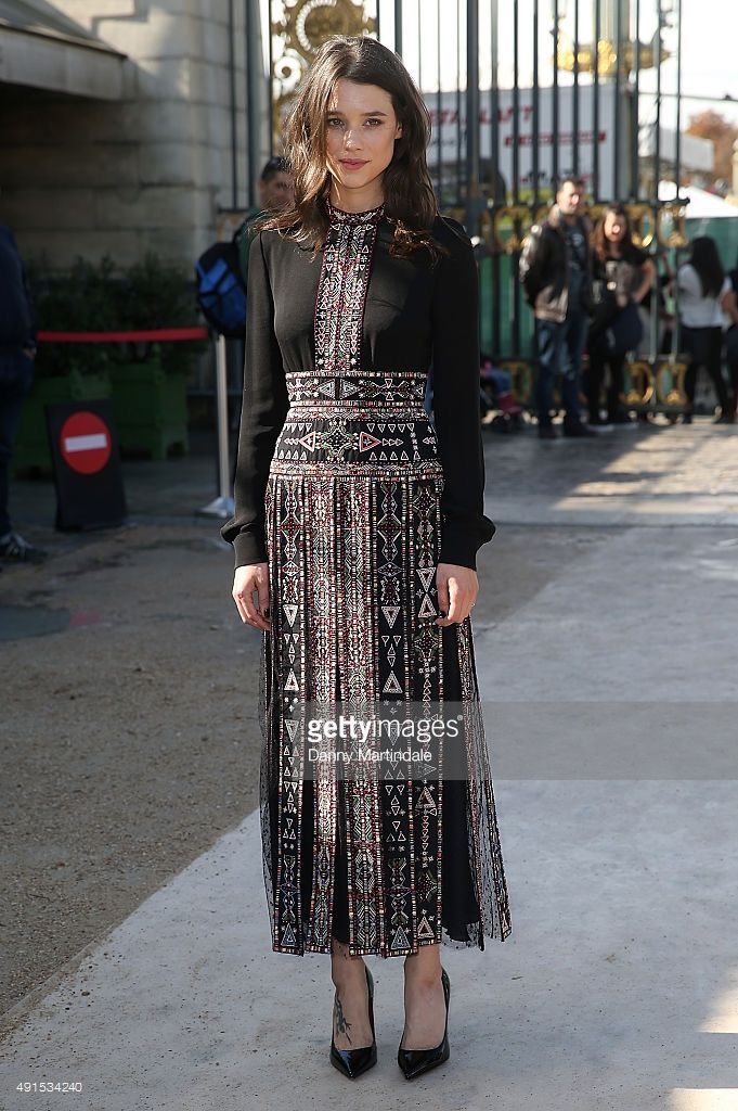 a woman is standing on the street in front of a gate. she is wearing a black dress with a pattern, and black shoes. the background includes a building and other people. there is a "getty images" watermark and the photographer's name, "danny martindale."