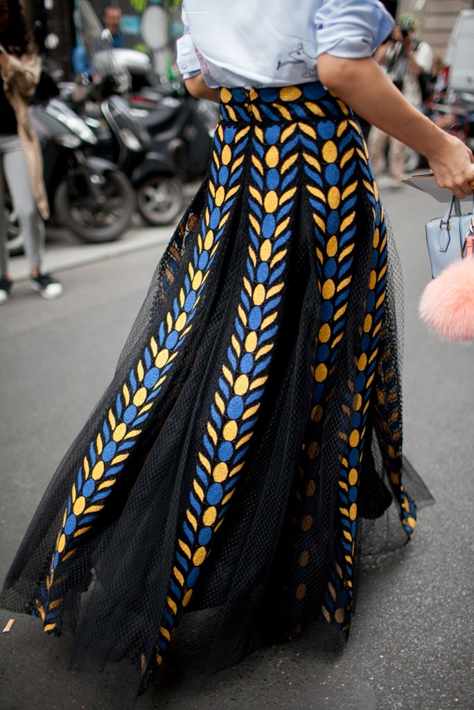 a person wearing a long black tulle skirt with a blue and yellow embroidered pattern on the sides is walking on a street. the person is also wearing a light blue top and carrying a light blue handbag with a pink pompom. the background shows a street scene with motorcycles and people.