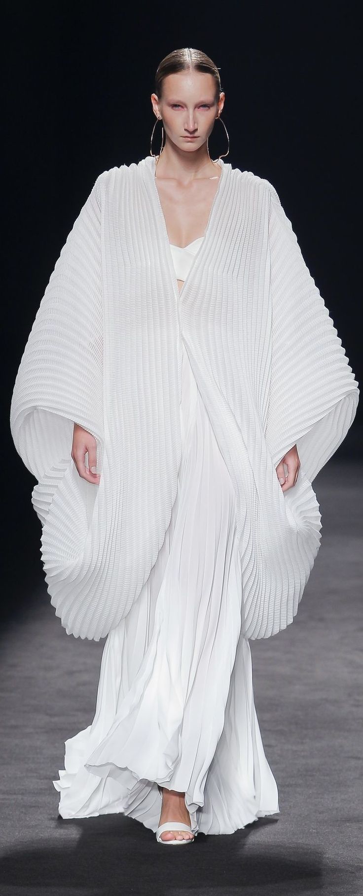 a model walks the runway during a fashion show, wearing a long, white, pleated gown and robe. she is wearing large hoop earrings and white sandals. the background is black.