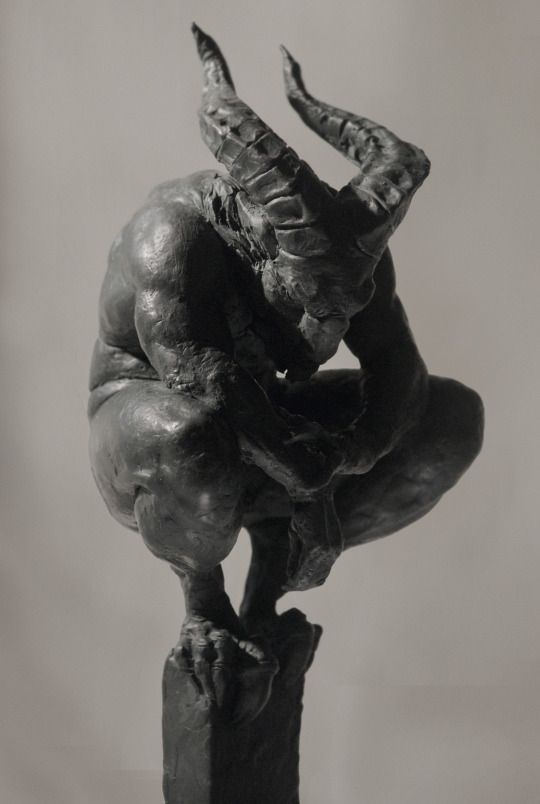 a black and white photo depicts a sculpture of a muscular, crouching demon with large horns. the sculpture is made of clay and placed on a pedestal.