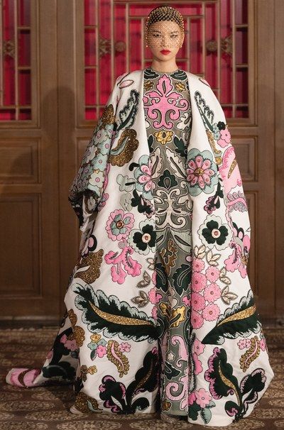 the image showcases a model wearing a stunning haute couture gown. the dress is a floorlength masterpiece, featuring a long, flowing cape design.  the fabric is predominantly white and is richly embroidered with an intricate floral pattern in shades of pink, green, and gold. the embroidery is highly detailed and looks heavily embellished. the model wears a delicate headpiece or veil, adding to the overall elegance of the look. the background suggests a luxurious setting, possibly a fashion show or photoshoot in a grand interior. the overall aesthetic is one of high fashion, sophistication, and intricate artistry.
