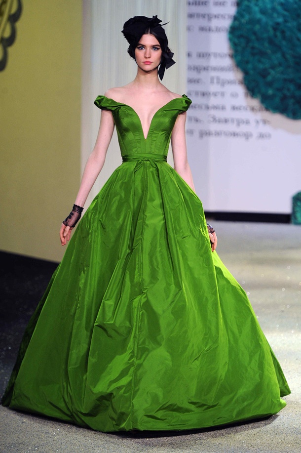 a fashion model wearing a voluminous, bright green offtheshoulder ball gown on a runway. she has a black headpiece and sheer black gloves. the background is a blurred fashion show setting with script visible on a screen.