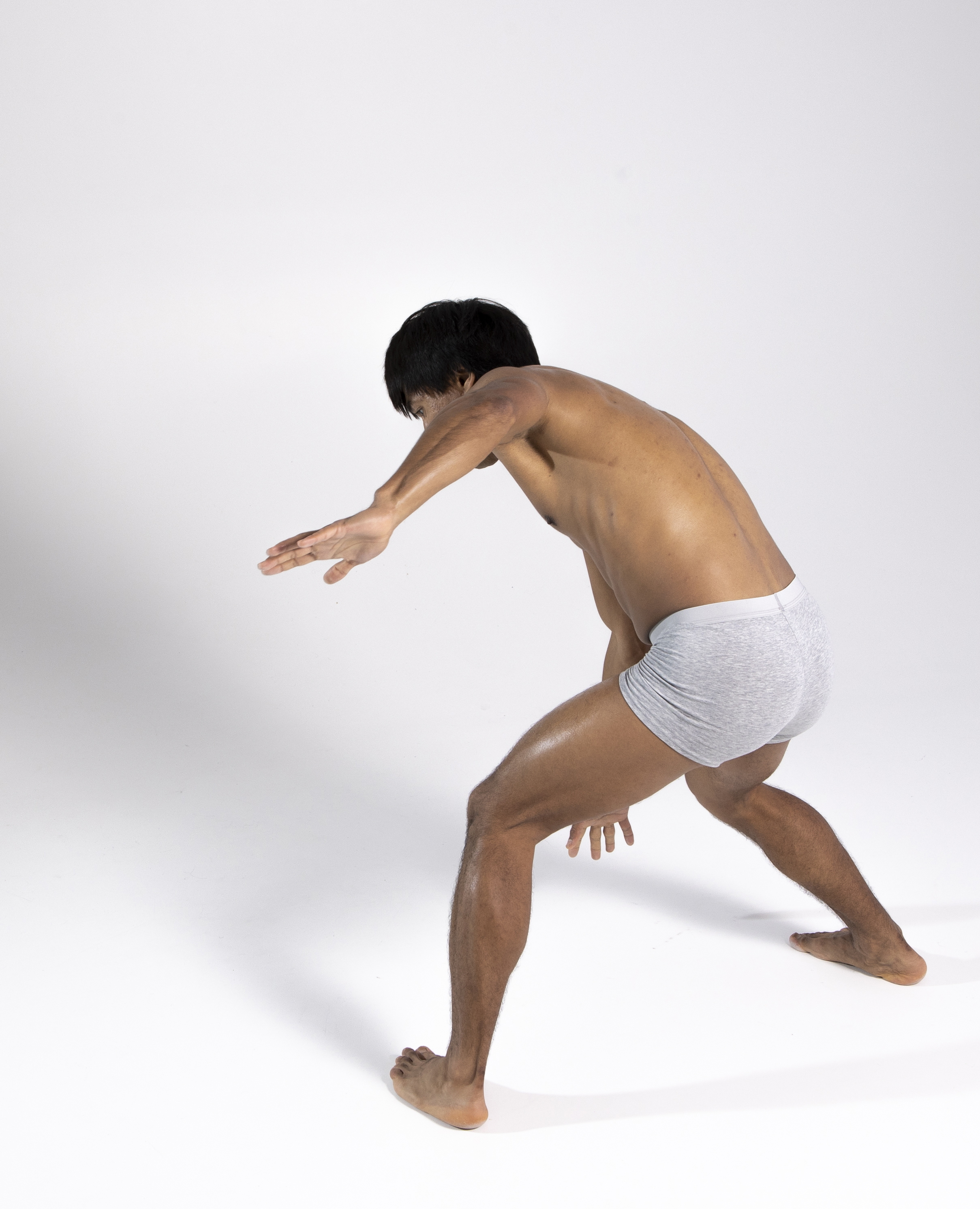 a studio shot of a barefoot, bare chested, adult, dark hair male dressed in underpants, bending with one hand reaching out, looking down.