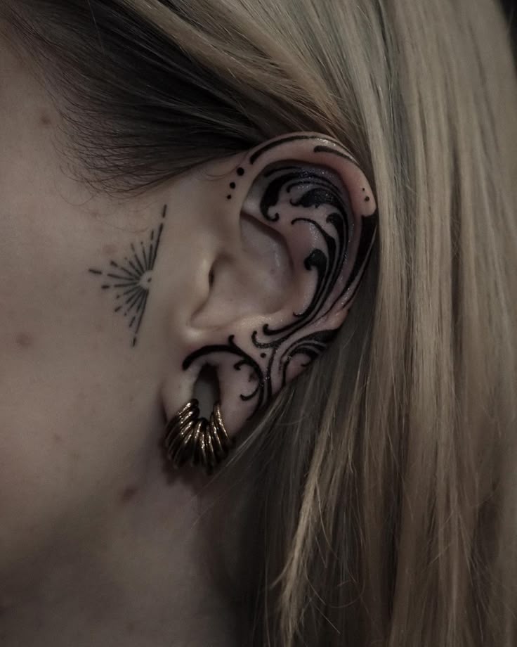 a closeup image of a person's ear with various tattoos and piercings. the ear is adorned with a large, black, wavelike tattoo that covers most of the outer ear. there are also a few dots tattooed near the top of the ear. the earlobe is stretched and contains multiple golden hoop earrings. on the cheek near the ear, there is a small, black tattoo of a sun. the person has blonde hair that falls around their face.