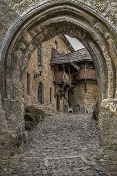 the image shows a stone archway leading into a medieval courtyard paved with cobblestones. beyond the archway, there is a building with a stone facade and a wooden balcony structure supported by stairs. a lantern hangs on the wall of the building. there are windows and doorways visible in the building. the overall atmosphere is historical and rustic.