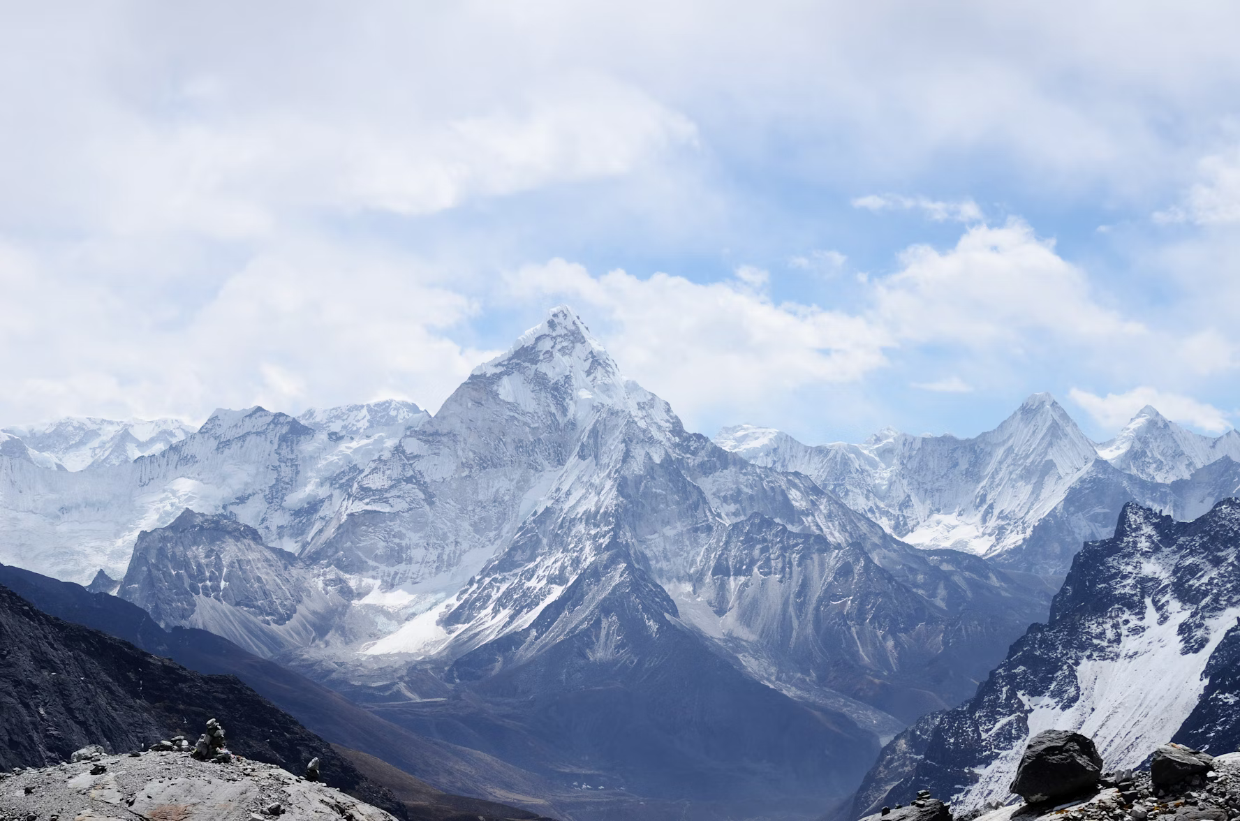 the image shows a panoramic view of a majestic, snowcapped mountain range against a backdrop of a blue sky dotted with fluffy white clouds. the mountains are predominantly white due to the snow cover, with shades of gray and brown visible where the rock is exposed.  the foreground suggests a high vantage point, perhaps another mountain's slope, hinting at the vastness of the himalayas.