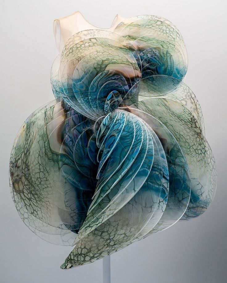 a closeup view of a stunning abstract glass sculpture that resembles a dress. the sculpture is made of multiple layers of glass that are intricately arranged to create a swirling, flowing design. the glass is painted in shades of blue, green, and white, giving the sculpture a sense of depth and movement. the sculpture is lit from the side, casting dramatic shadows that enhance its beauty. the background is a soft gray.