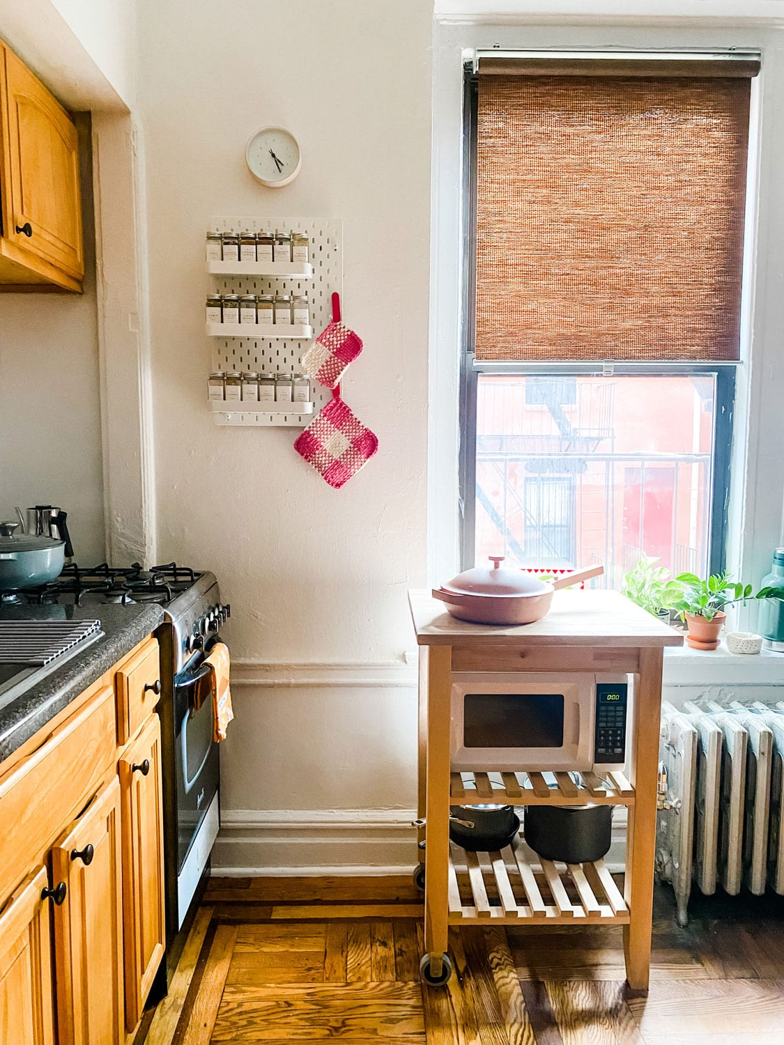 his is a photo of a small galley kitchen in what appears to be a new york city apartment. the kitchen has wood cabinets, wood floors, and white walls. there is a window on the right side of the image with a woven bamboo shade. a microwave is on a wooden cart with wheels in front of the window. to the left of the microwave cart is a white radiator cover. next to the radiator cover is a plant in a terracotta pot on a wooden stand. on the wall to the left of the microwave cart is a spice rack and a pink and white crocheted pot holder. the cabinets on the left side of the image have black countertops and a stainlesssteel stovetop. there is a gray pot on the stovetop.  the walls and cabinets are white. the image is brightly lit with natural light coming from the window.