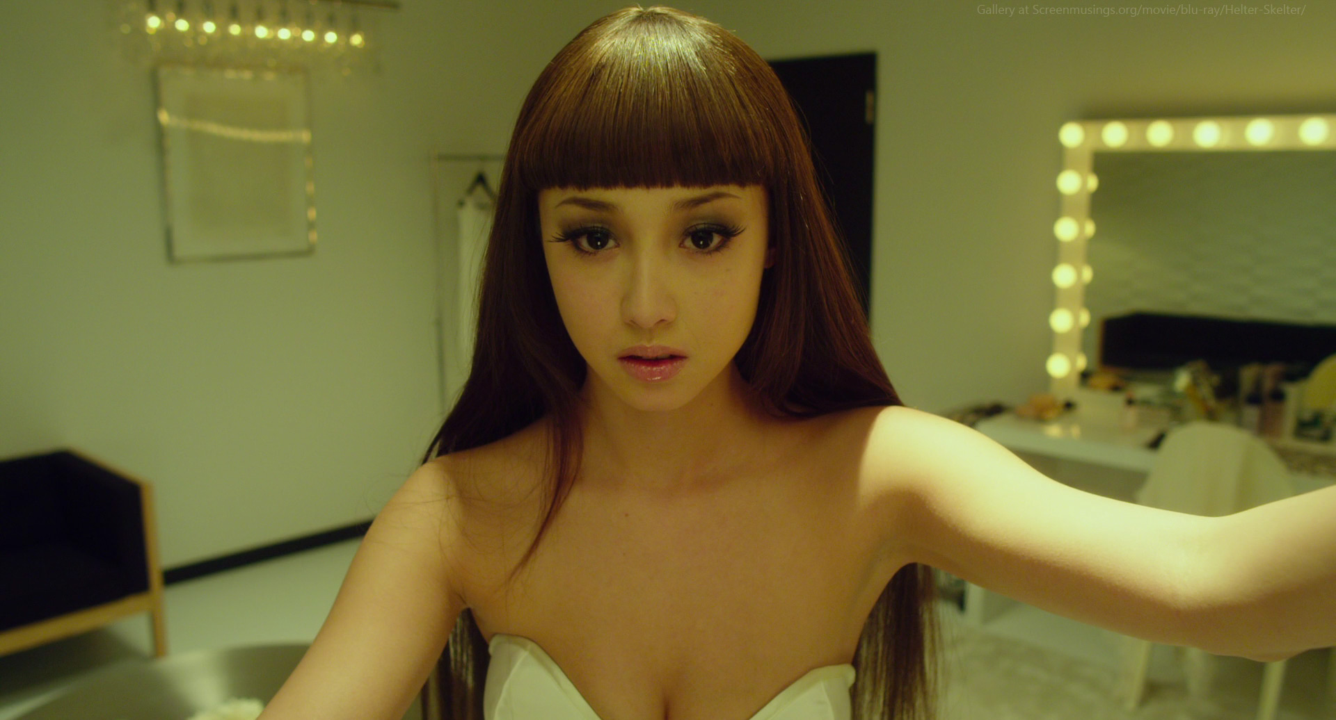 a young woman with long, dark hair and a blunt fringe takes a selfie in a welllit dressing room. she is wearing a strapless white dress and has dramatic eye makeup. the background features a mirror with vanity lights, a white dressing table, and a wooden chair.