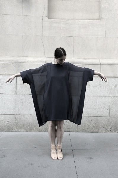 a woman is standing in front of a wall, posing in a dark, oversized dress and lightcolored shoes. her arms are outstretched.