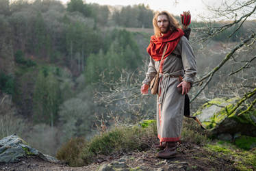 a man dressed in medieval attire stands on a rocky hillside in a forest. he is wearing a long grey robe with a red hood or scarf, brown boots, and a leather belt and pouch.  he carries a bow and quiver suggesting he's an archer. the background shows a lush green forest. the overall mood is serious and historical, suggesting a scene from a historical drama or fantasy setting.