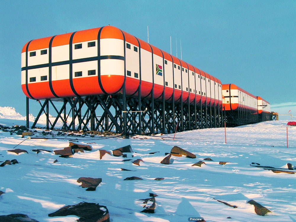a long red, white, and black building with a south african flag sits on stilts in a snow covered landscape. the building is most likely a research station in antarctica.