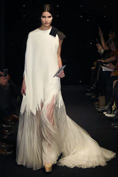 a fashion model in a white long gown is walking on a runway. the dress has a sheer overlay and a delicate design. she is looking straight ahead and poses with a hand holding an accessory in front of her. the model's hair is styled in a simple, elegant manner and she is wearing minimal makeup. the background is dark and blurry, creating a stark contrast with the model's white gown. the overall effect is one of elegance and sophistication.