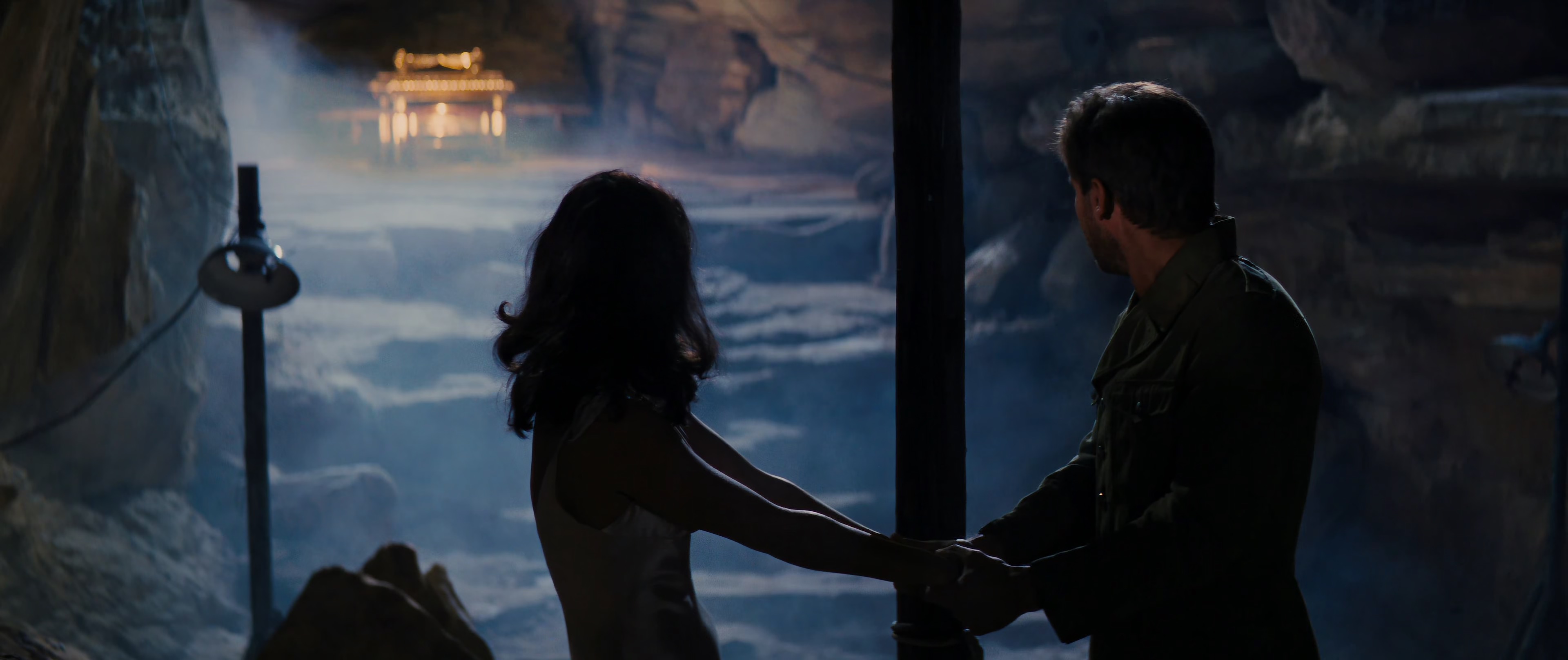 a couple stands in a dark cave, holding hands. the man is on the right, wearing a military jacket, while the woman is on the left. they are both silhouetted against the light of a temple in the distance. the scene is dramatic and mysterious, with the couple's love and commitment shining through in the darkness. the background is filled with rocks and fog, adding to the mystical atmosphere.