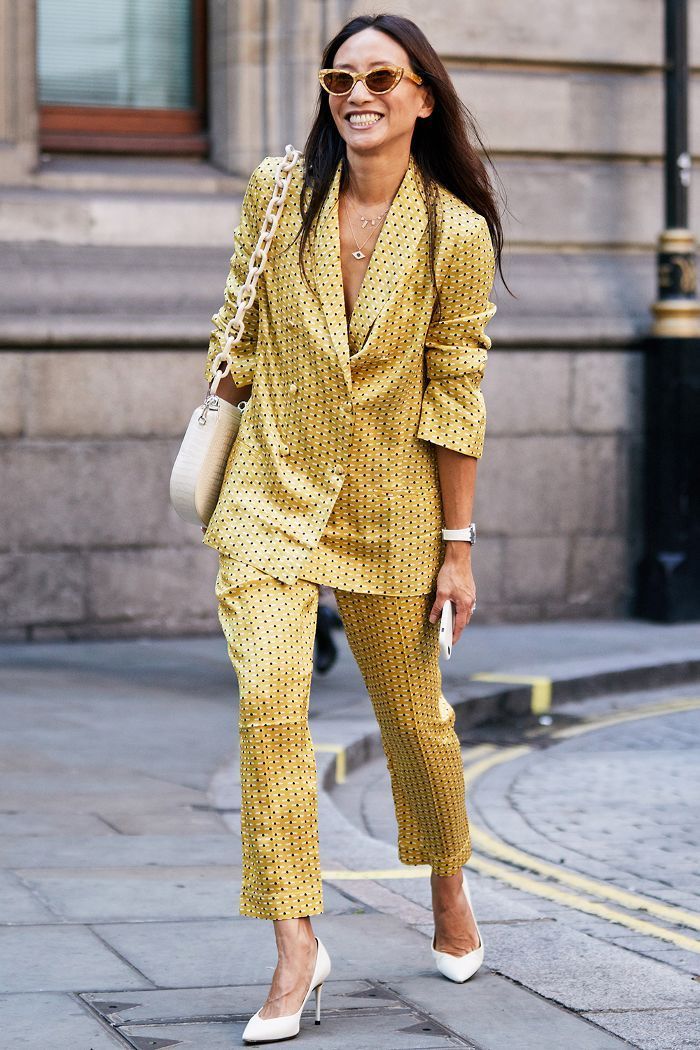 a stylish woman in a yellow polka dot pantsuit strolls down a city sidewalk. she's wearing white heels, sunglasses, and a chain bag. her confident smile and trendy outfit make a statement of summer fashion. the image is taken outdoors in natural light.