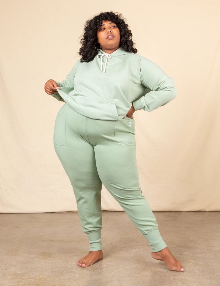 a plus size woman in a green hoodie and matching sweatpants stands barefoot against a light beige background. she has curly hair and is posed casually with her arms crossed at her waist. she's looking toward her right side of the frame.