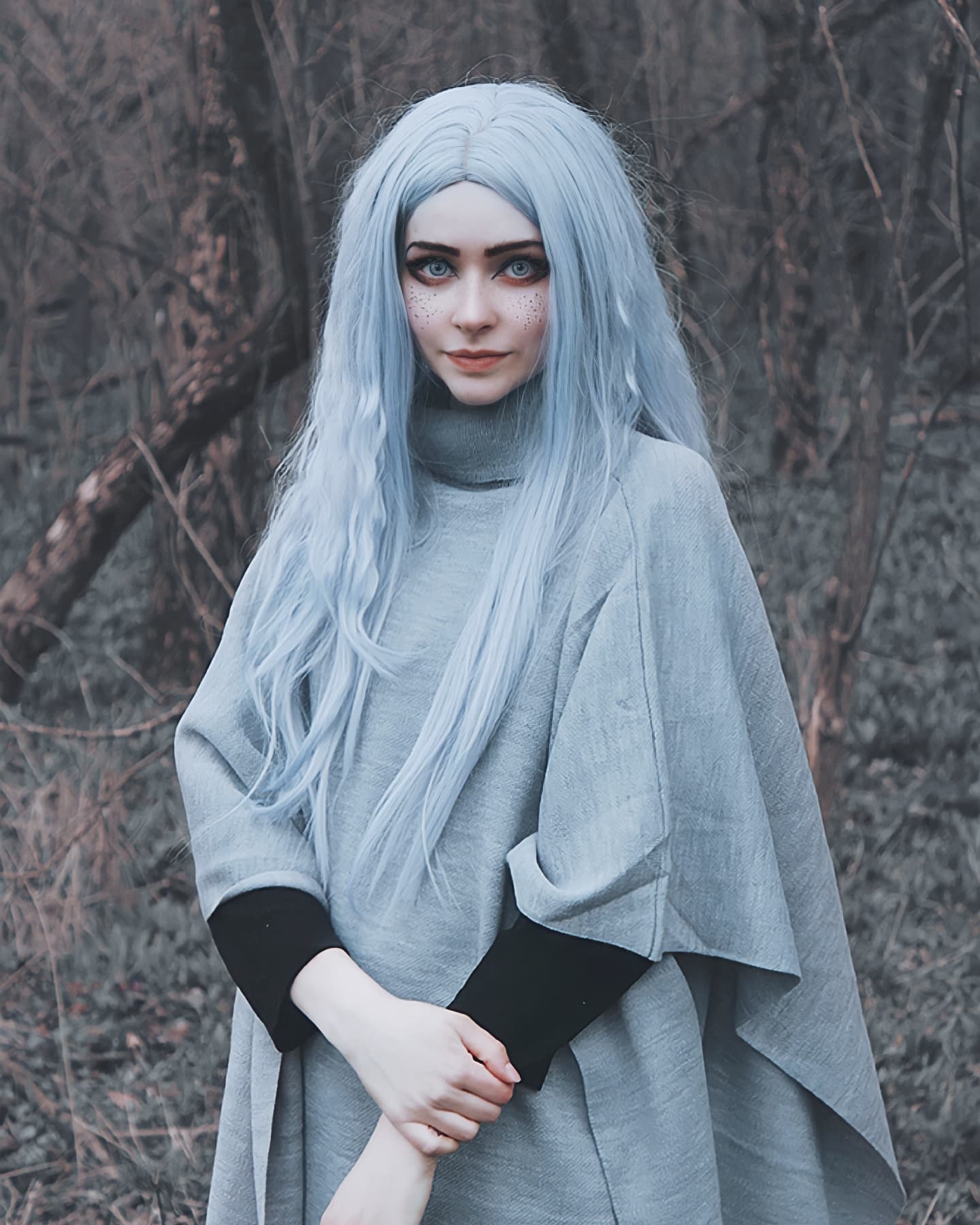 the image depicts a young woman with long, pale blue hair styled in loose waves. she's wearing a light grey poncho or cape with black cuffs and a highnecked, turtleneckstyle top underneath. her skin is exceptionally pale, almost white, and she has subtle freckles across her cheeks. the overall aesthetic is mystical and ethereal, reminiscent of an elf or other fantasy character.  she stands in a dark, somewhat shadowy forest setting, with bare, leafless trees visible in the background. the woman looks directly at the camera with a calm and serene expression. the lighting is soft and diffuse, enhancing the overall mystical feel of the photo.