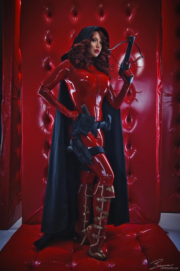 a woman in a red latex superhero costume with a cape and boots is holding a crossbow. she is standing in a red room with a red couch. she is looking at the camera.