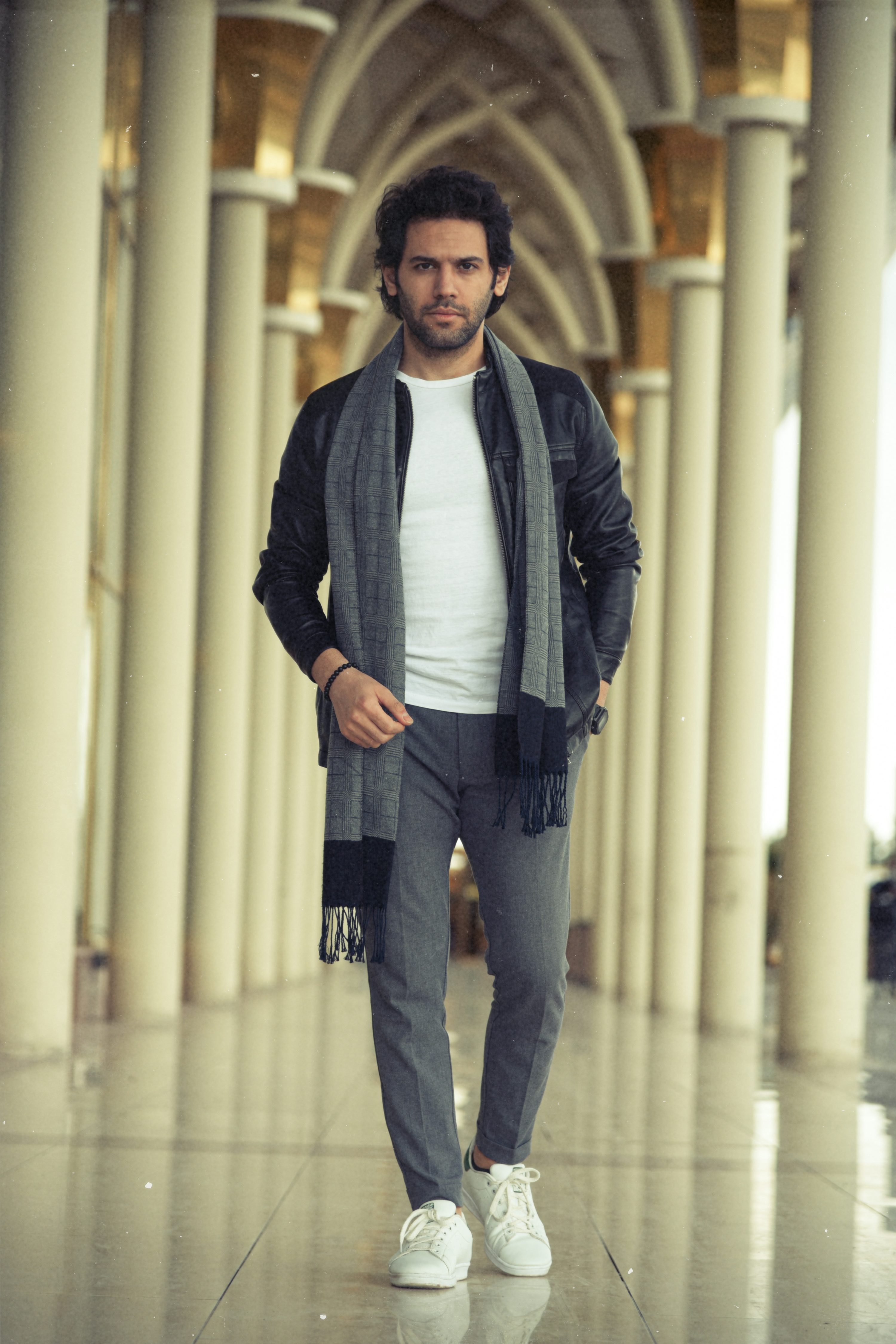 a full shot of a stylish man walking down a hallway with columns. he is wearing a grey scarf, a black leather jacket, grey pants, and white sneakers. his expression is serious and he is looking directly at the camera. the background is blurry but shows the architecture of the building. the overall aesthetic is trendy and fashionable.