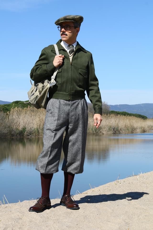 a man dressed in vintage clothing stands by a river. he's wearing a green jacket, grey tweed knickerbockers, burgundy shoes, and a newsboy cap. he carries a canvas bag over his shoulder. the setting is outdoors with a tranquil river and sandy bank.  the scene evokes a retro or vintage feel, possibly 1930s or 1940s style.  the sunlight indicates a daytime setting, likely springtime, based on the lack of heavy foliage.