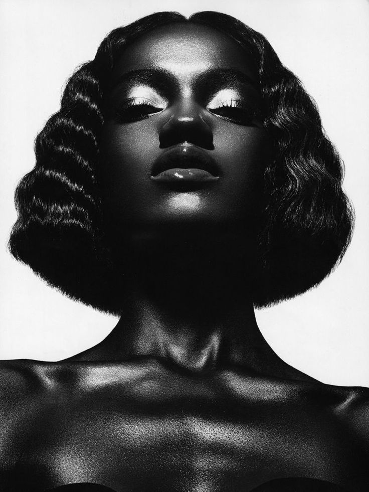 the image is a striking black and white closeup portrait of a darkskinned woman. she has a short, wavy bob haircut and her eyes are closed, giving off a serene and almost meditative expression.  the lighting is dramatic, creating strong highlights and shadows that emphasize her features and the texture of her skin. her shoulders and neck are visible, framing the portrait. the overall style is elegant and sensual, suggestive of a fashion or beauty campaign.