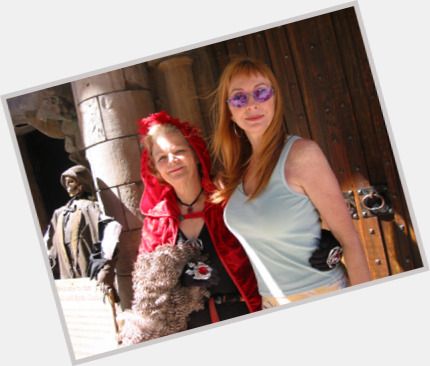 two women are posing for a photo. the woman on the left is wearing a red cloak and has curly hair, while the woman on the right is wearing a blue top and has long, straight hair. they are standing in front of a doorway. the woman on the right has her hand on the shoulder of the woman on the left, and they are both smiling.