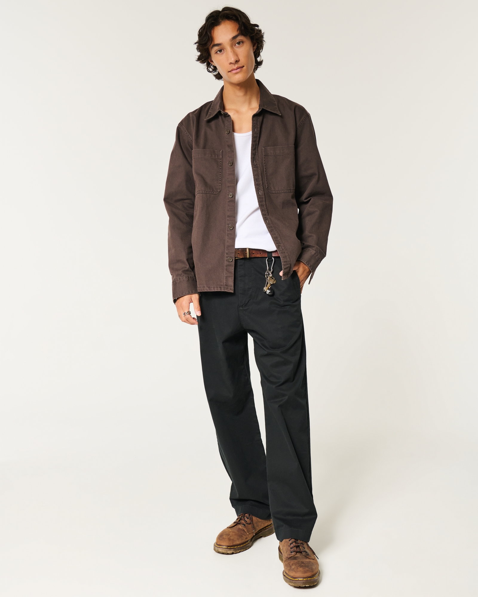 a fullbody studio shot of a male model showcasing a casual outfit. he's wearing a dark brown longsleeved shirt, black wideleg trousers, and brown leather boots.  he has a white undershirt visible at the collar and a simple keychain hanging from his belt.  the background is plain white.