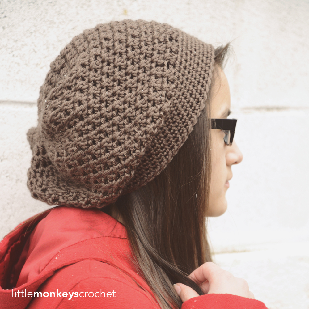 the image shows a young woman wearing a crocheted beanie hat. the beanie is a slouchy style, made from a textured brown yarn.  the crochet stitches are clearly visible, showcasing the intricate texture. the woman has long brown hair and is wearing a red jacket. the background is a white brick wall. the image is welllit and the focus is on the beanie and the woman's profile.  the watermark "littlemonkeyscrochet" is present in the bottom left corner, suggesting this is a design from that creator.
