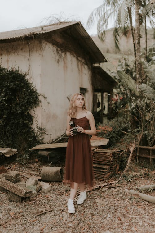 a young woman in a brown tulle midi dress and white sneakers stands in front of an old, abandoned house. she is holding a vintage camera and looking away. the setting is rustic and overgrown with plants and trees.