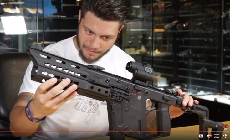 a man with a beard wearing a white tshirt is holding a black vector kriss rifle with an optic sight attached to it. he is indoors, sitting at a table, and behind him is a glass case containing a gun collection.