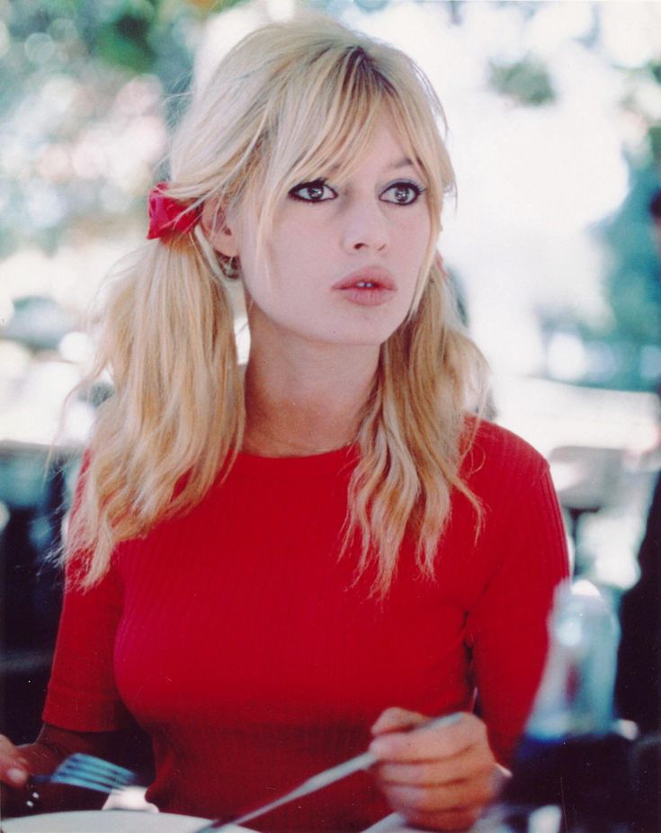a vintage photo of brigitte bardot, a famous french actress. she is sitting at a table outdoors, likely at a restaurant. she's wearing a red top and has her blonde hair styled in pigtails with red ribbons. she is looking offcamera with a contemplative expression and is holding a fork in her right hand, seemingly about to eat. the image is a closeup portrait with natural lighting.