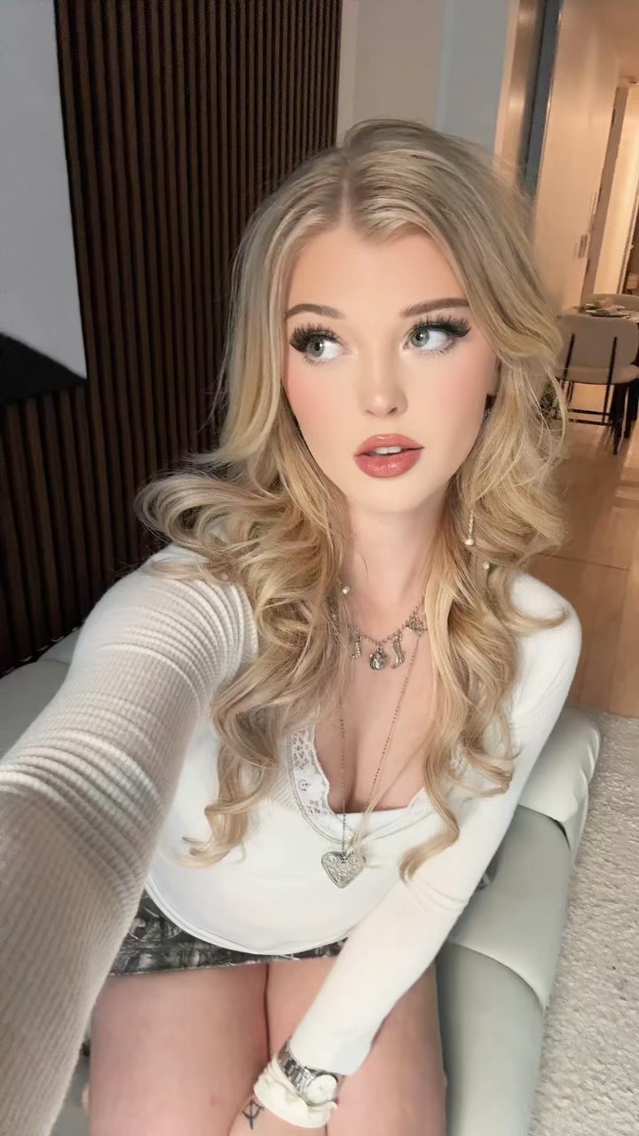 a woman with blonde hair is taking a selfie indoors. she is wearing a white long sleeve shirt and has multiple necklaces and a watch on. she also has a tattoo on her wrist.