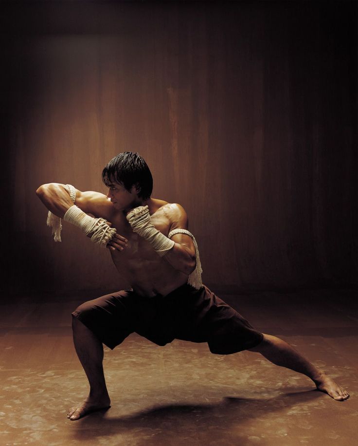 a man in traditional muay thai attire, wrapped in white fighting bandages on his arms and legs,  stands in a fighting stance.  he is in a darkened room with a textured brown background.  he is looking intently at a point off screen, as if he is shadow boxing.  the lighting is  dramatic and highlights his muscles and powerful body.  he is ready for action,  intense and focused, demonstrating the power and skill of the ancient art of muay thai.