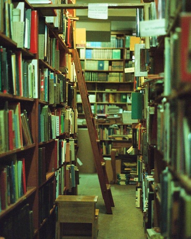 the image shows a narrow aisle between tall, wooden bookshelves packed with numerous books. a wooden ladder leans against one of the shelves, adding to the vintage feel. the lighting is dim, casting a green tint over the scene, creating a cozy and peaceful atmosphere. the perspective of the photo leads the viewer down the aisle, emphasizing the depth and vast quantity of books in the library. a small wooden stool sits near the bottom of the image, adding to the quiet setting. the overall impression is one of a classic, wellloved library filled with a multitude of books.